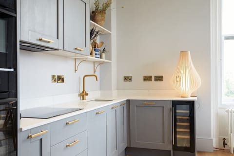 Modern kitchen featuring soft gray cabinets, gold fixtures, and built‑in wine cooler. Open shelving with curated décor and sculptural lighting blend elegance with guest‑friendly comfort.