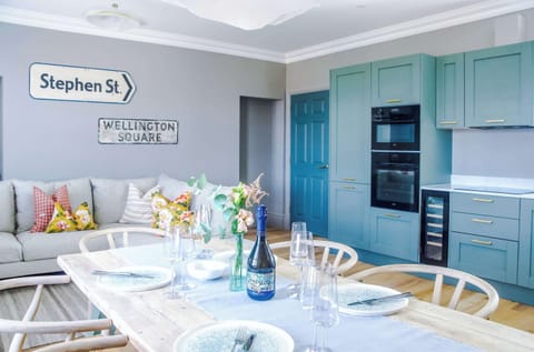 Contemporary living room with a plush beige sofa and patterned cushions, accented by quirky street-sign décor. Warm lighting and cloud-like fixtures add charm, creating a cozy, stylish space that flows into the kitchen and dining area.