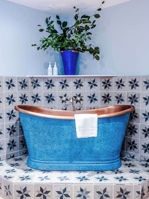 Stylish bathroom with a freestanding blue tub and copper rim set against patterned tiles. A recessed shelf with greenery, soft lighting, and a gold-framed mirror add elegance, while natural light creates a cozy, inviting retreat.
