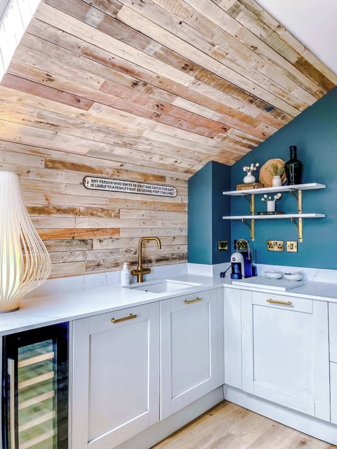 Modern kitchen corner with slanted wooden ceiling, white cabinetry, and gold accents. A glowing sculptural lamp and floating shelves with decor add warmth, while muted green walls and sleek finishes create a stylish, inviting space.
