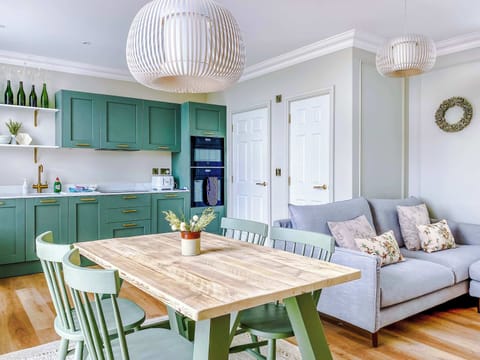 Sage cabinets, white counters, and built‑in oven set a refined tone. Wooden dining table, pendant lights, and cozy lounge seating enhance the inviting atmosphere, making the space both functional and stylish