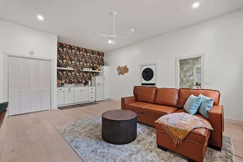 Living area with full kitchen view. Cognac leather sectional with throw pillows, louvered closet doors, in-unit washer/dryer, and full kitchenette with designer wallpaper accent.