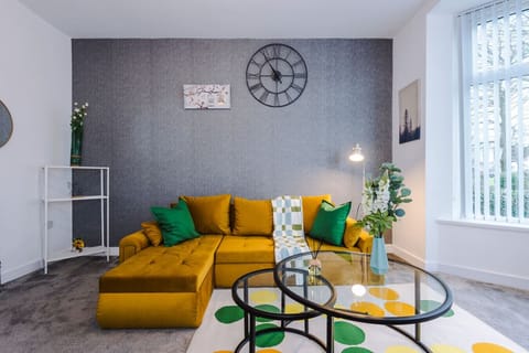 Living room with a mustard corner sofa, round glass coffee table, wall clock, and a window with vertical blinds.