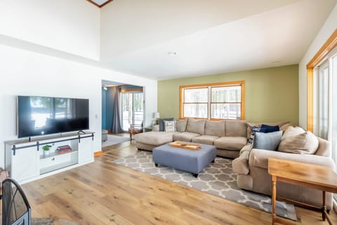 Living Room - Comfy couch, smart TV, and fireplace. Off to the left is the piano room, to the right is a walkout to the back porch. The camera POV is from the base of the stairs to the upper bedrooms and game room, and off to the left is the entry to the kitchen.