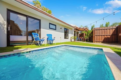 Sunny sleek pool, blue lounge chairs, and a stainless steel grill. String lights and tropical plants add charm, while artificial grass and a tall wooden fence create a private, stylish space for outdoor relaxation.