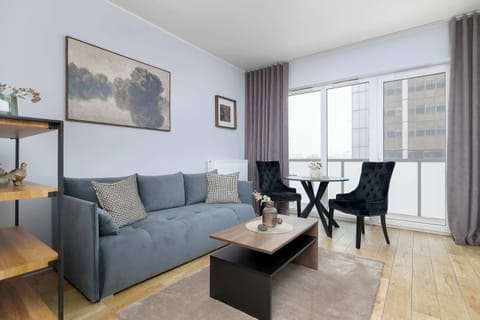 A comfortable seating area featuring a gray sofa and a small dining table placed near large windows. Natural light fills the room, complemented by long gray curtains. The layout connects the lounge space with a compact dining corner.