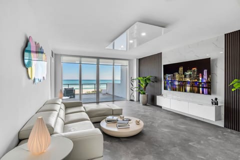 Relax in a chic, minimalist living area with a plush sectional and floor-to-ceiling glass doors opening to ocean views. This modern space features marble accents, sleek wood paneling, and a large flat-screen for ultimate coastal luxury.