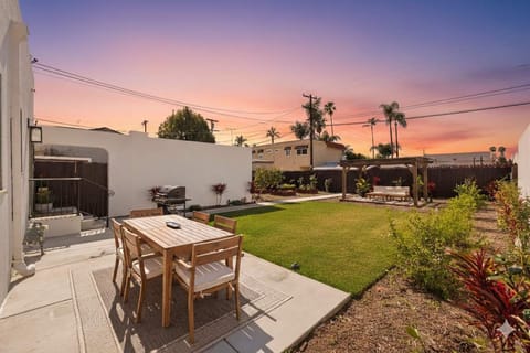 Enjoy a beautiful sunset in this spacious backyard featuring a manicured lawn, wooden dining set, and a cozy pergola. Perfect for outdoor entertaining under a clear blue sky.