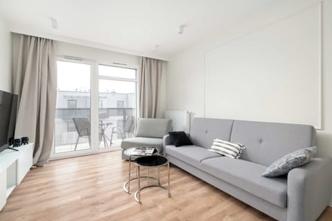 The living room area opens up to a balcony through large glass doors, blending indoor and outdoor spaces. A stylish grey sofa and a circular glass coffee table sit on light-colored laminate flooring.