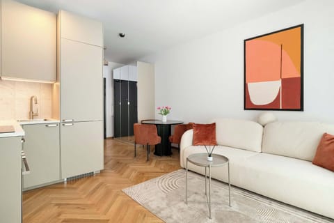 A wide-angle view of the living room and kitchen area, highlighting the cream sofa, coffee table, and compact white kitchenette along the back wall. Bright natural light enhances the clean design.