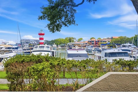 Verdant hedges frame a vibrant view of the marina, where yachts bob gently under a bright azure sky. The iconic red and white lighthouse stands as a colorful sentinel over this peaceful waterfront scene.
