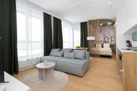 Bright studio interior with a comfortable grey sofa, large windows dressed in deep green curtains, and warm wooden flooring creating a welcoming and elegant atmosphere.