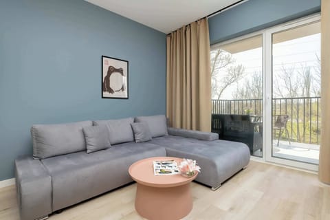 A living room view featuring a gray sofa positioned near a large sliding glass door. Natural light pours in, and greenery is visible outside. A small round coffee table stands in front of the sofa.