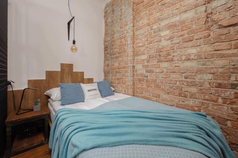 A bedroom with an exposed red brick wall, a wooden headboard, and a neatly made bed covered with a turquoise blanket and matching pillows. Soft lighting hangs from the ceiling, adding a cozy industrial feel.