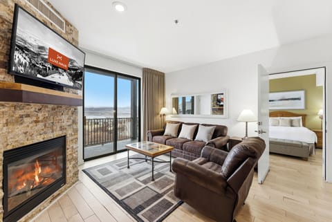 Main Living Area with Fireplace and Balcony:
Open-concept living room with leather sofa, stone fireplace, large windows, and private balcony offering mountain views.