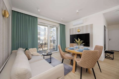 A bright living space features a comfortable beige sofa, a round wooden dining table with four elegant chairs, and a large window with  curtains.