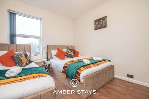 Two beds with neutral headboards are accented by vibrant orange and teal linens in this bright, minimalist room. Warm wood flooring and cream walls complete the clean, inviting look.