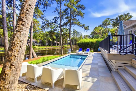 Shimmering pool waters reflect the afternoon glow, surrounded by smooth stone tiling and towering pines. This quiet backyard escape borders a peaceful canal, offering a private spot to lounge under the vast blue sky.