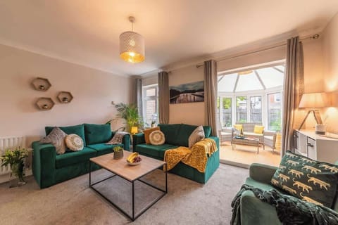 Living area with sofa bed, 2 seater sofa and chair in plush green. There is a door leading to the conservatory and garden. TV present on opposite wall at Blue Puffin Stays Short Lets & Serviced Accommodation