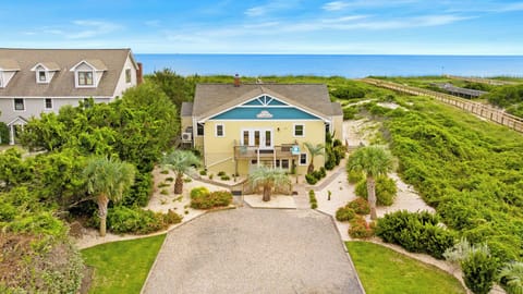 Oceanfront Oak Island NC beach cottage with yellow siding, blue trim, covered entry, upper deck, gravel driveway, palm trees, lush dunes, and private boardwalk leading directly to the Atlantic Ocean.