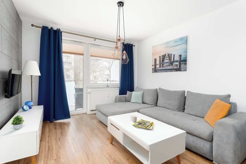 A bright living room with a gray corner sofa and a white coffee table placed on wooden flooring. Blue curtains frame a large window that lets in natural light. A flat-screen TV stands on a low white media unit against the wall.