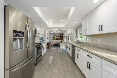 Bright contemporary kitchen with stainless steel appliances, elegant white cabinetry, and light stone countertops seamlessly flowing into a cozy living area for the perfect blend of style and functionality.