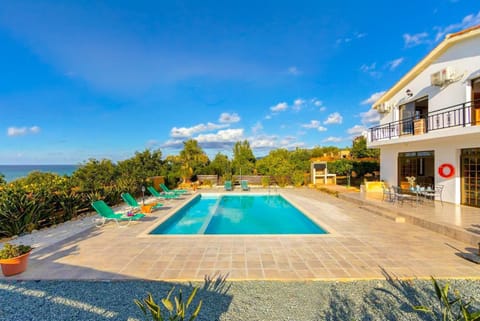 Large private swimming pool with turquoise water on a tiled patio, green sun loungers, an outdoor dining table, and a two-story white villa overlooking the ocean under a clear blue sky.
