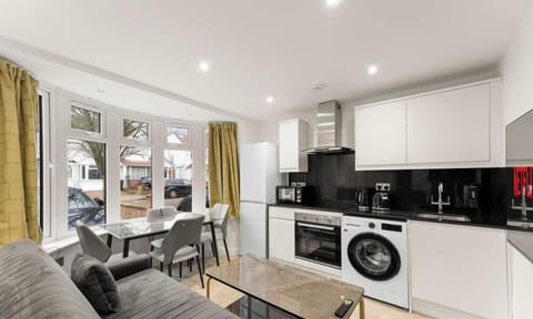 This compact living space features a modern white kitchenette and a grey sofa positioned near a bright bay window. Mustard-yellow curtains add a pop of color to the otherwise neutral, sunlit room.