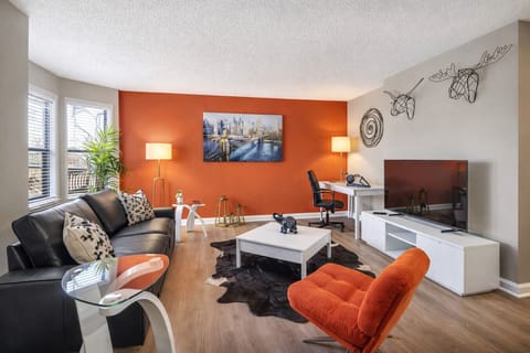 Bright, modern living room with a bold orange accent wall, plush leather sofa, and stylish décor. Enjoy a 55” Smart TV, dedicated workspace, and natural light—perfect for relaxing or working comfortably in the heart of Uptown Charlotte