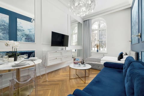 An elegant living room with high ceilings, white walls, and large arched windows that flood the space with natural light. A deep blue velvet sofa contrasts beautifully with the bright interior. A round white coffee table and a wall-mounted TV complet