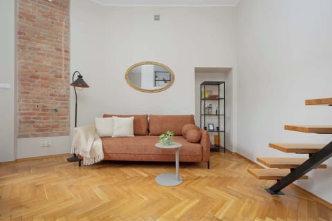 Bright studio living area with a cozy sofa, exposed brick wall and wooden stairs leading to the mezzanine level.
