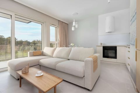 A bright, open-plan living area features a comfortable L-shaped cream sofa facing a wooden coffee table. The space is seamlessly connected to a modern kitchenette and a dining zone near large windows.