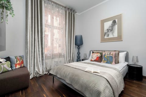 A bright studio apartment with a large bed placed beside tall windows and soft curtains. The open layout combines sleeping and living space into one airy room.