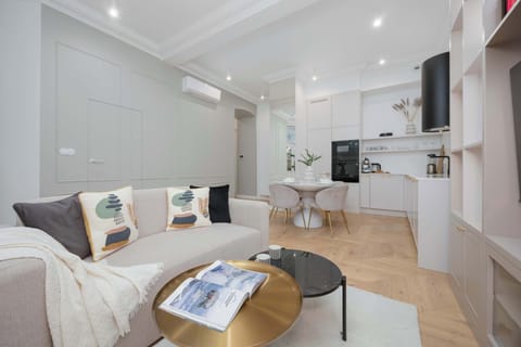 A bright living room with a sofa, coffee table, and open kitchen in the background. Light colors and built-in shelving give the space an airy feel. Decorative pillows add warmth to the seating area.