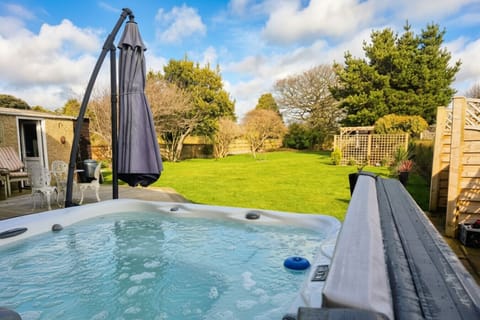 A white hot tub filled with water sits on a paved patio in front of a large lawn, with a garden umbrella, outdoor chairs and timber fencing visible beyond.