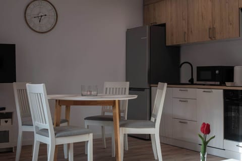 A bright dining area featuring a round wooden table with four white chairs, situated next to a modern kitchenette. In the background, a grey refrigerator and minimalist cabinets are visible, with a red tulip on the table adding a pop of color.
