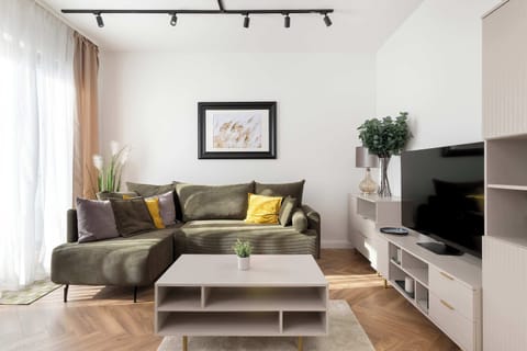 The focal point of the lounge is a comfortable, tufted green sofa facing a modern white media console with a large TV. A minimalist coffee table sits on a light-colored rug, creating a cozy relaxation zone.