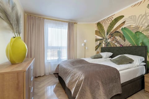A cozy bedroom features a large double bed with plush pillows and a botanical-themed wallpaper depicting lush tropical leaves. A wooden dresser stands to the left, topped with a vibrant yellow decorative vase that adds a pop of color to the room.