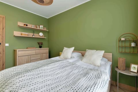 A tranquil bedroom displays a large double bed with crisp white linens and a decorative green accent wall. Wooden floating shelves and a matching dresser add a touch of natural warmth to the interior.