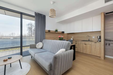 A bright living area with a gray sofa, round coffee table, and floor-to-ceiling windows. Light wood cabinetry and neutral tones create a modern, airy atmosphere.