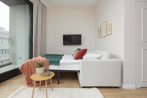 A living space featuring a white sofa and wall-mounted TV near the entrance door. Simple artwork decorates the wall. The room appears tidy and contemporary.