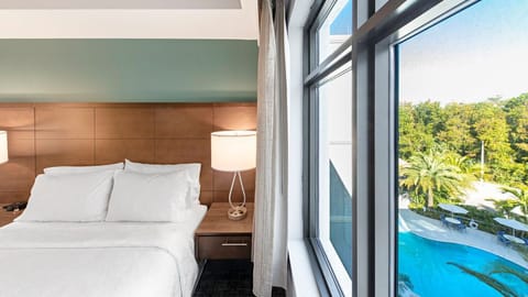 The room features a queen bed, bedside lamp, and a large window overlooking the outdoor pool and palm trees.