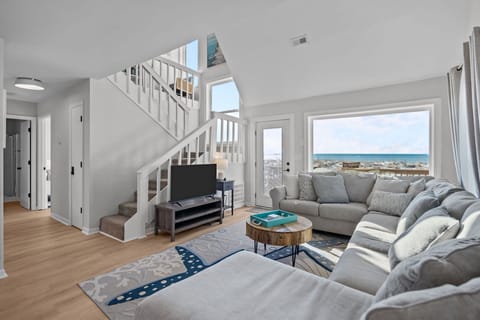 Bright oceanfront living room in Oak Island NC with large sectional sofa, starfish area rug, round coffee table, TV, coastal artwork, and direct deck access with beautiful Atlantic views through oversized windows.