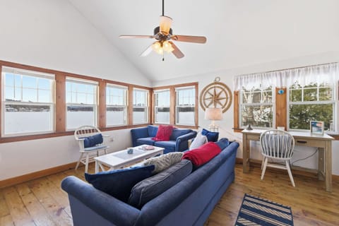 The lake light fills this open living room all day, with two blue sofas, pine floors, and a nautical wheel on the wall keeping things grounded in classic New England cottage character.