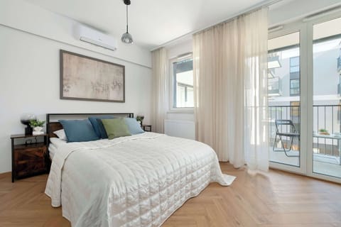 A spacious bedroom space features a large, made-up bed as the centerpiece. There's also a window on the right side of the photo and a piece of art on the left wall.