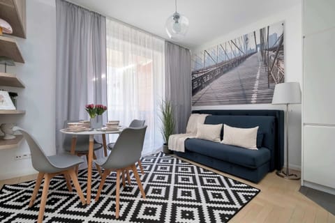A bright living room connected to the dining room, where the blue sofa and stylish rug with geometric patterns catch the eye. A round table with two gray chairs has been placed by the window, creating a cozy coffee corner.