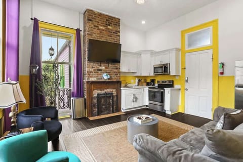 Open-concept living area with comfortable seating, exposed brick fireplace, and Smart TV, flowing into a fully equipped kitchen with modern appliances. Bright, colorful space that blends historic character with everyday comfort.