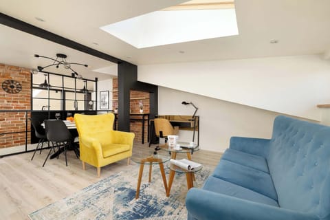 A bright open-plan living area features a blue sofa and a striking yellow armchair set against white sloped ceilings. The space is illuminated by a large skylight and decorated with a patterned rug and industrial-style lighting.