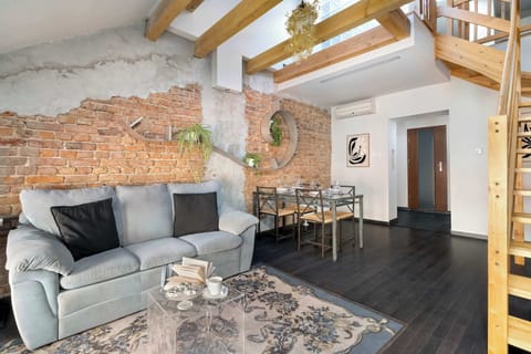 A cozy living room with a gray sofa and exposed brick wall, accented by wooden beams and decorative plants. A patterned rug and soft lighting create a warm, inviting atmosphere.