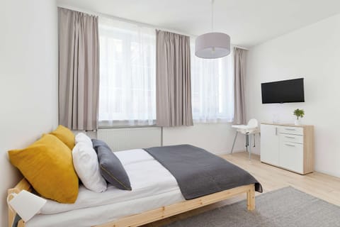 A bright and tidy studio apartment bedroom area features a single bed with gray and yellow pillows, a white curtained window, a dresser, and a wall-mounted television.